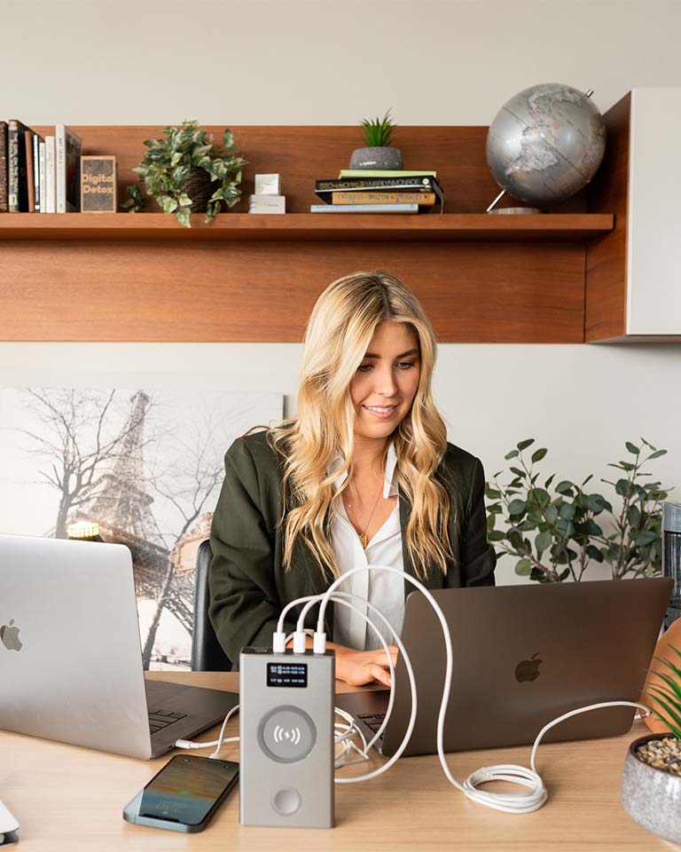 Image of female using Graphene Powerbank, Flash Pro Plus to charge 2 x Macbooks and iPhone 14. 25000mAh large battery capacity, OLED display, USB-C 100W fast charging. An iPhone 14 is being charged on the MagSafe compatible wireless charging up to 15W. An Apple Watch is being charged on the separate Apple Watch charging pad supporting up to 5W. Perfect for Apple users on the go. 